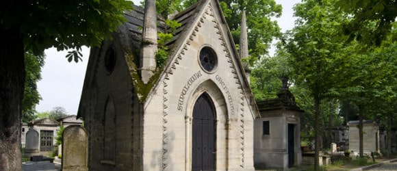 Custom-built stone mausoleum with pointed roof and arched door, nestled among trees and cobblestone path in serene cemetery setting. Forever Legacy premium craftsmanship and concierge service in the USA.