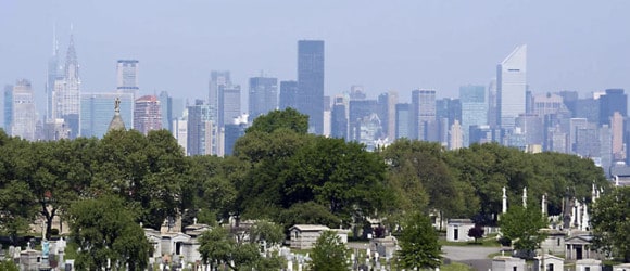 Cemetery with headstones in foreground; city skyline and tall buildings in background. Custom private mausoleums by Forever Legacy - expert craftsmanship, concierge service, perpetual guarantee throughout the U.S.