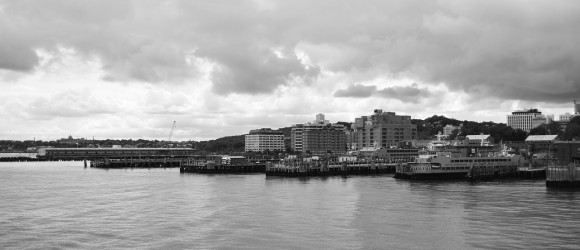Black and white image of waterfront with elegant mausoleums by Forever Legacy, featuring buildings, ferry, and dramatic cloudy sky backdrop. Premium custom-built private mausoleum services for high-end clients in the USA.