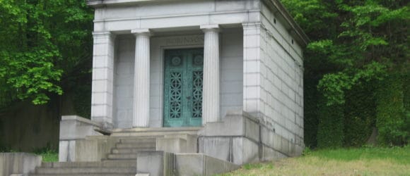 Gray stone mausoleum with four columns, green doors; surrounded by lush trees and grass. Elegant private mausoleums by Forever Legacy.