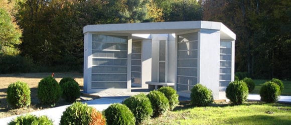 Luxury custom-built mausoleum by Forever Legacy, nestled amidst shrubs and a concrete pathway in a serene park-like setting with tree-lined backdrop. Expert craftsmanship and concierge-level service guaranteed.