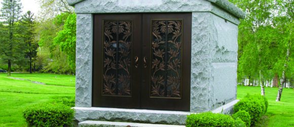 Ornate stone mausoleum with metal doors in a verdant cemetery, crafted by Forever Legacy for luxury private memorials.